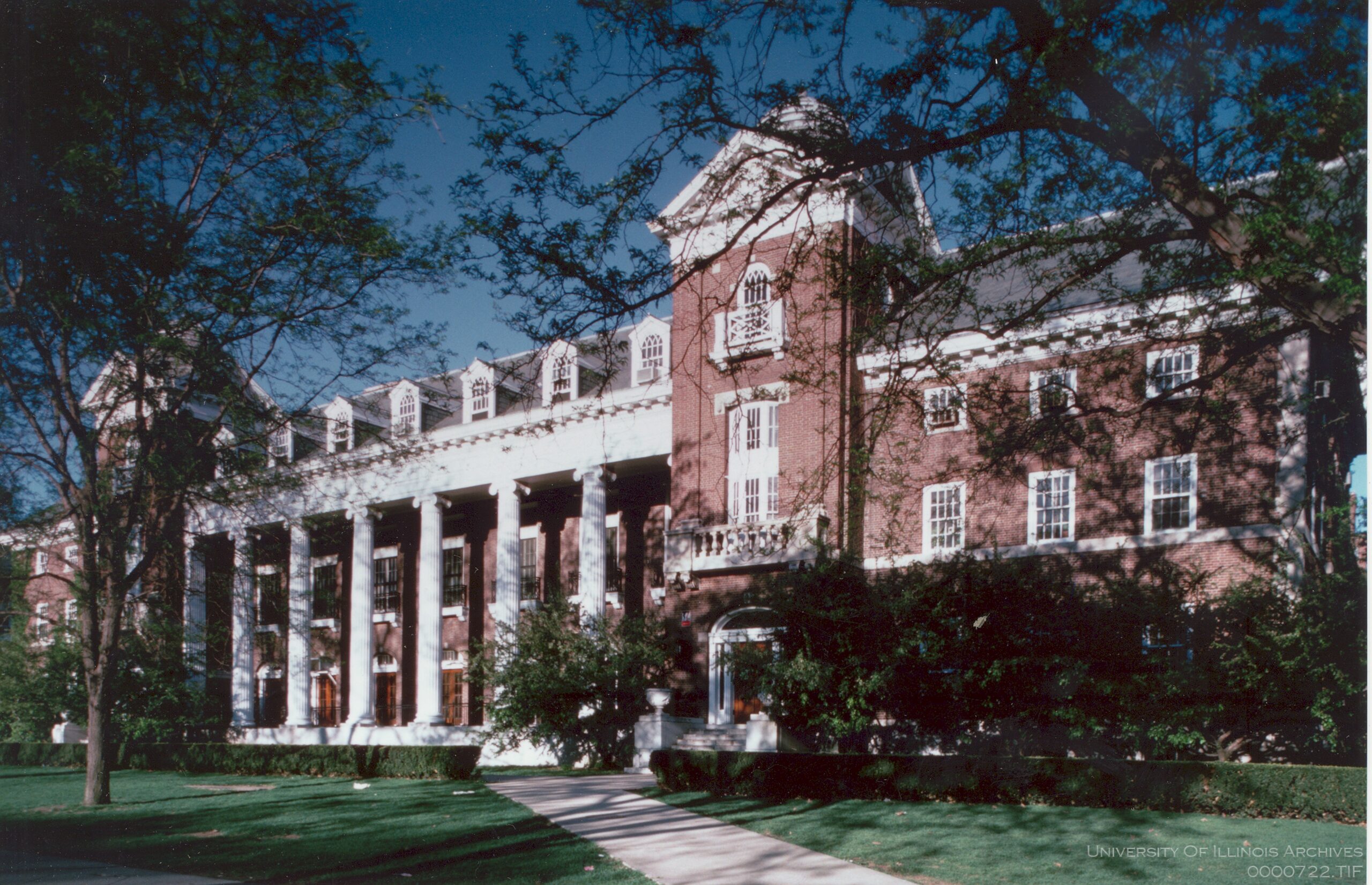 Color photograph of the University of Illinois English Building from northeast on a clear sunny day. Tall leafy trees foreground a big red brick building with white columns and window trim.