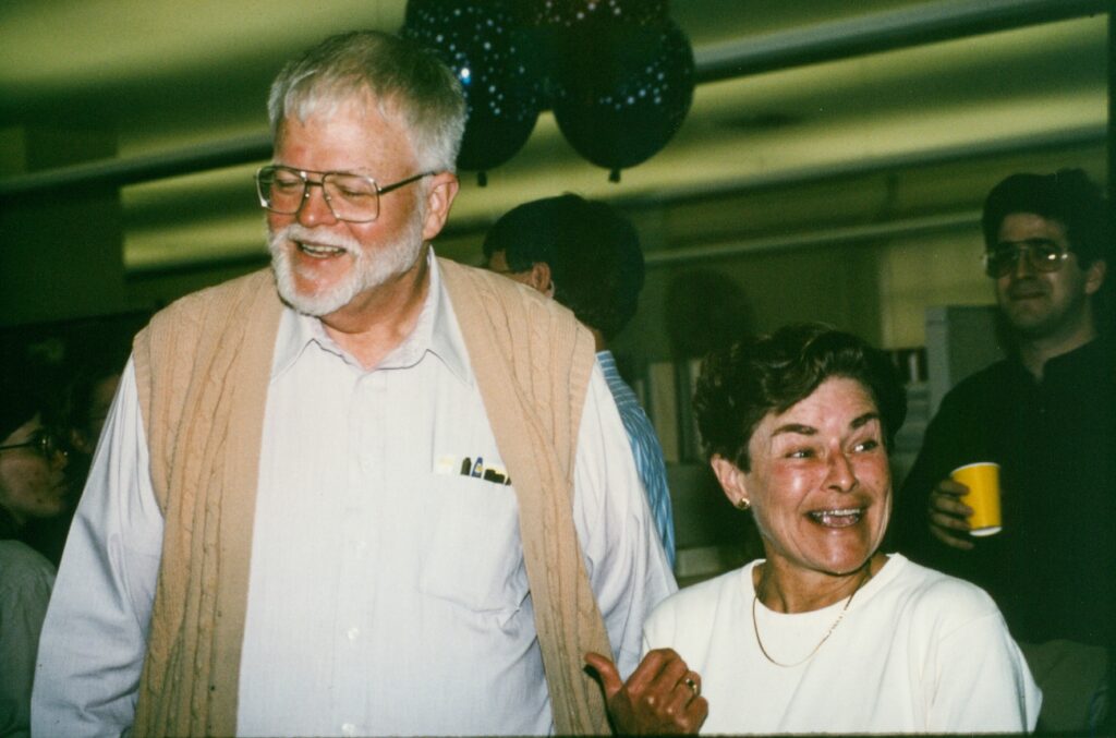 a smiling man and woman of very different heights face toward a camera, the woman gestures at the man with her thumb