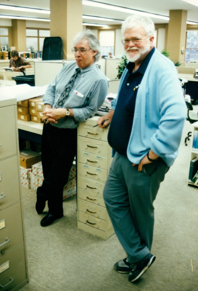 two men stand on either side of a beige filing cabinet, one in a shirt and tie, the other in a baby blue cardigan and sneakers