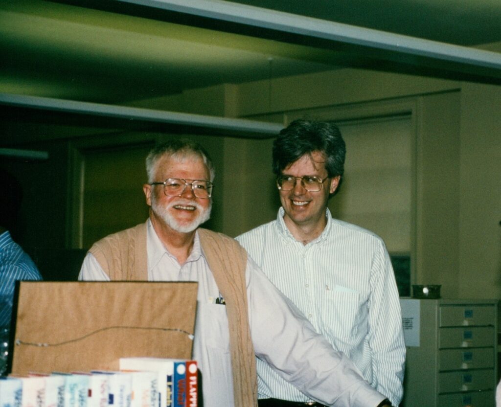 two men standing in an office space, both smiling, one smiling at the other