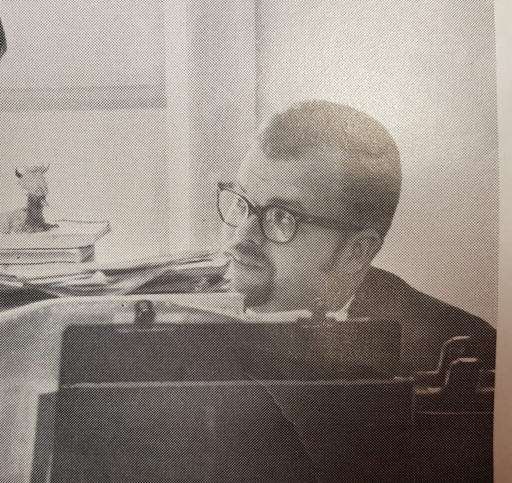 Young man in glasses looks to his right from behind a stack of papers, folders, and books.