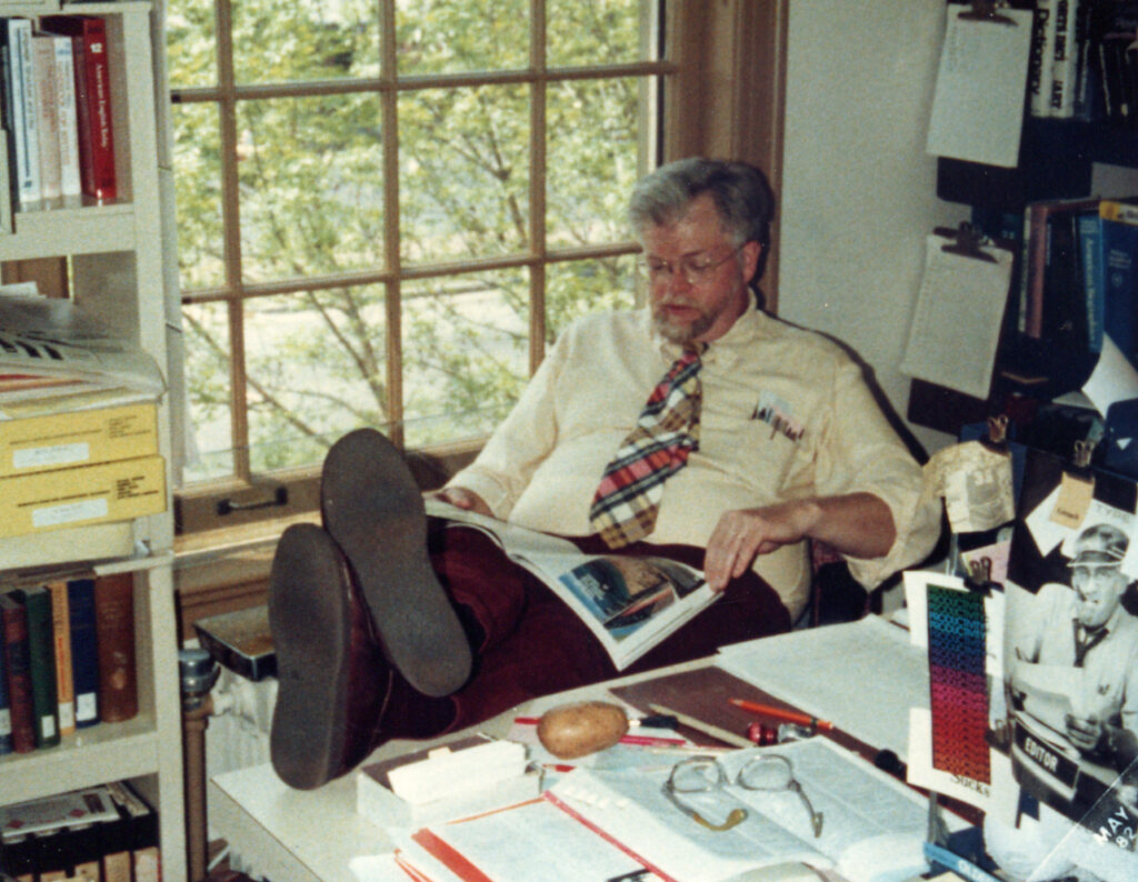 Man with glasses sits reading in front of a window, his legs up and crossed on a desk crowded with paperwork and a potato
