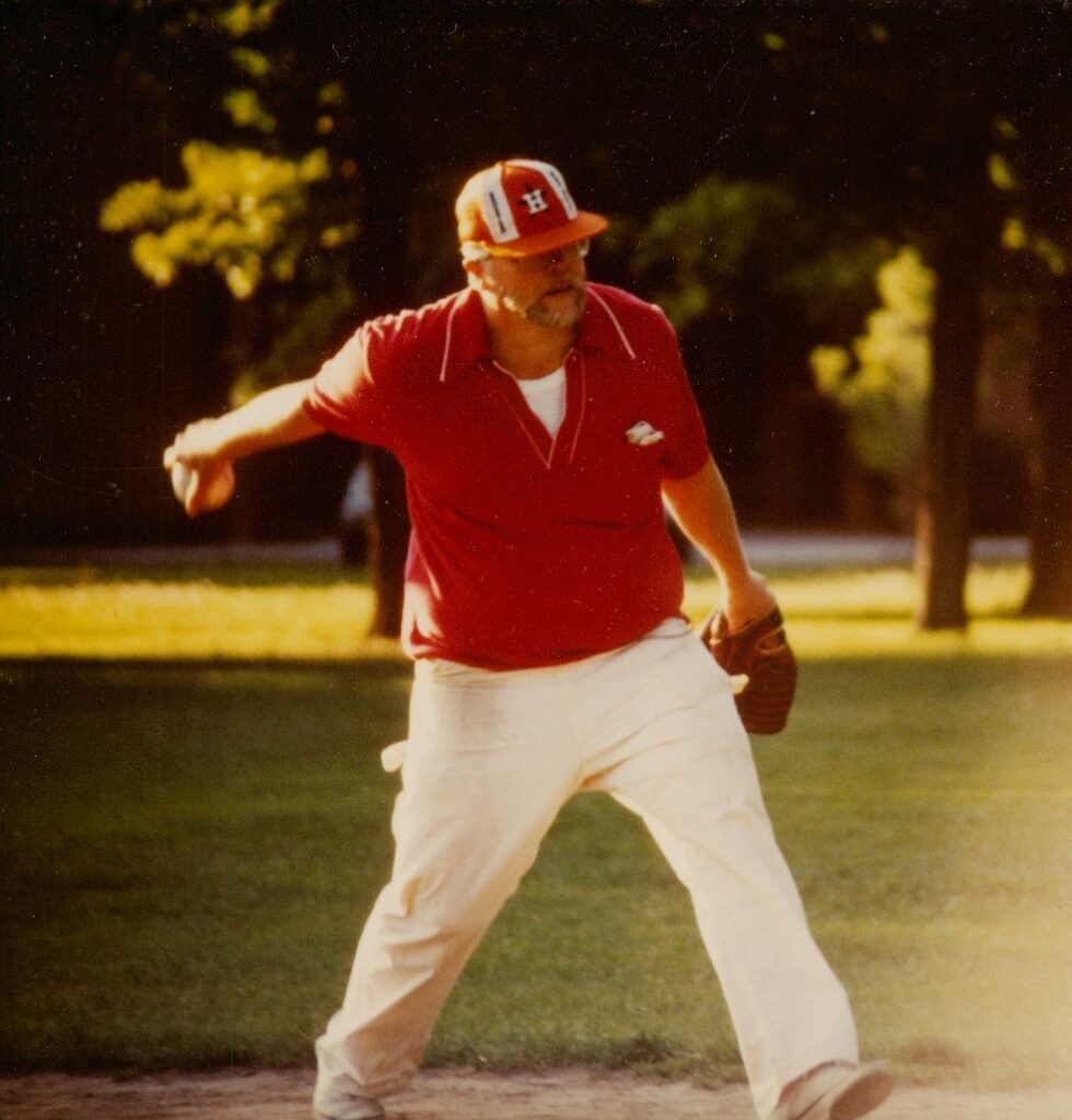 a man in a red shirt and baseball cap throws an underhanded pitch on a sunny green field