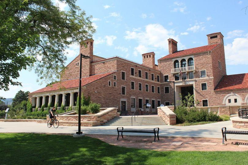 sandstone building surrounded by a bicyclist, benches, cafe tables, bushes, trees, and blue sky.