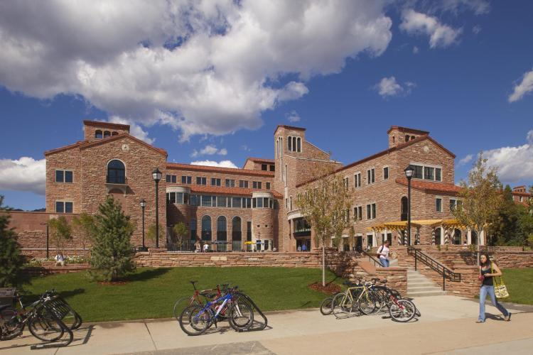 Sandstone building against a blue sky with fluffy white clouds. Rows of bicycles in the foreground, students walking.