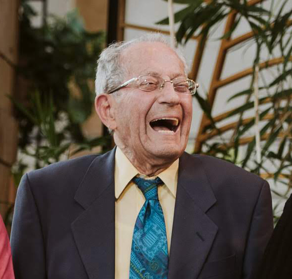 Ari Kernerman laughing. He's a man with grey hair, a blue patterned tie with a pale yellow shirt and dark blazer, pictured in front of greenery.