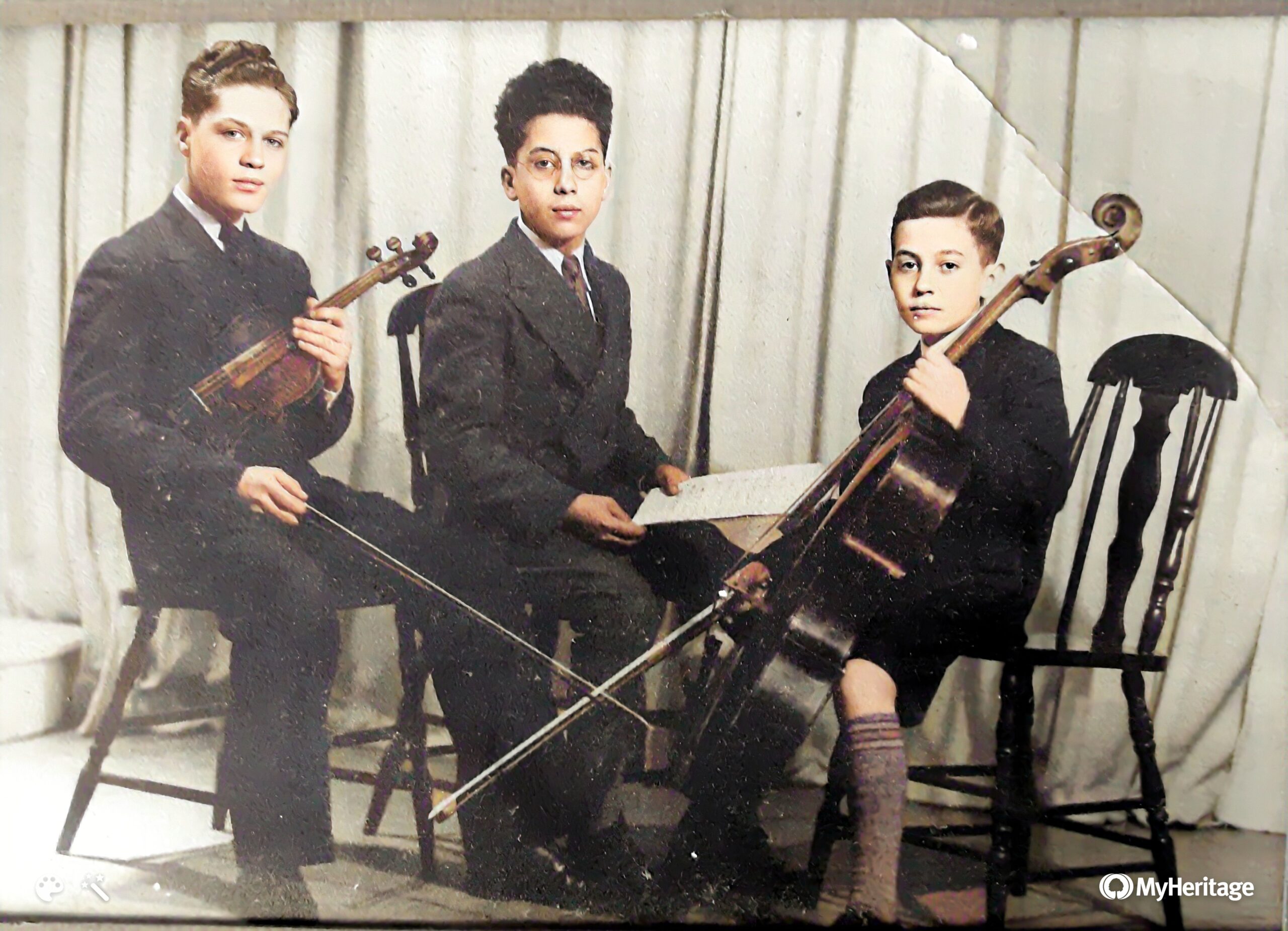 Three boys in dark suits seated before a white curtain. The boy on the left holds a violin, the boy in the center sheet music, the boy on the right a cello.
