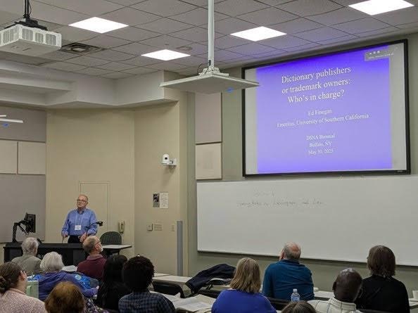 Ed Finegan in a blue shirt and white name badge stands at a podium in the lower lefthand corner. A projection at the center of the image reads "Dictionary publishers or trademark ovwers: Who's in charge?"