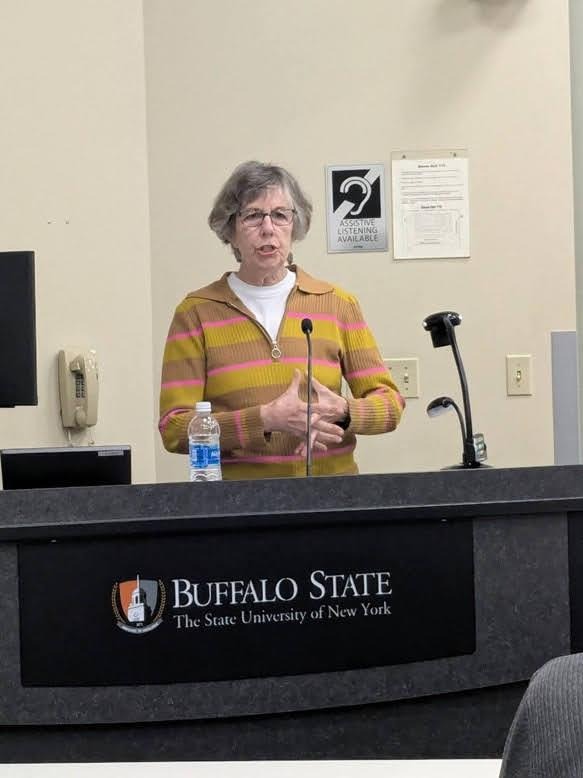 Karin Michaelson in a white shirt and cheerful sweater speaks from a podium  emblazoned with Buffalo State's logo.