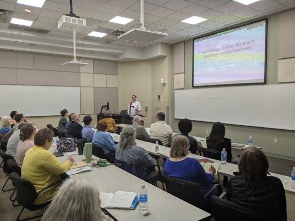Christian DiCanio in a white shirt with a red stripe speaks from a podium at the center of the image, a projection to the right of him is a powerpoint slide that reads "Constructing a Triqui dictionary: challenges and perspectives." Three rows of audience members are seated at long white tables in a room full of beige paneling.