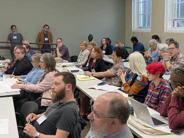 About thirty humans, wearing name badges and seated at beige tables or leaning on taupe walls, look ahead or take notes.