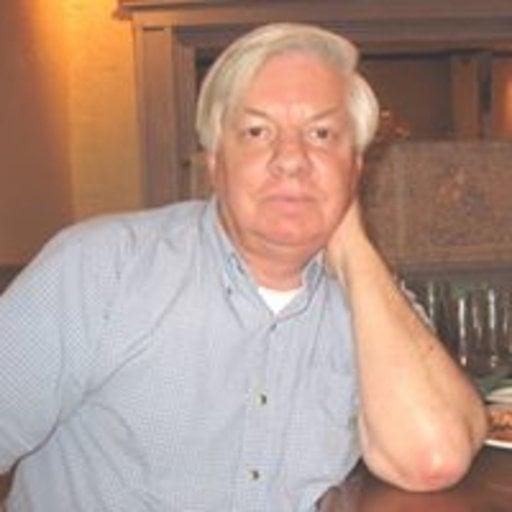 Ken Litowski, a white man with lush white hair parted at the left, brown eyes, and sharp eyebrows, wears a short-sleeved blue check button up and leans on his left arm propped on a table. The background is warm oranges and browns.